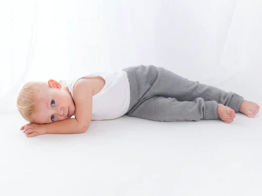 Young child with blond hair lying on their side on a white surface, wearing a white tank top and gray pants, looking calmly toward the camera.