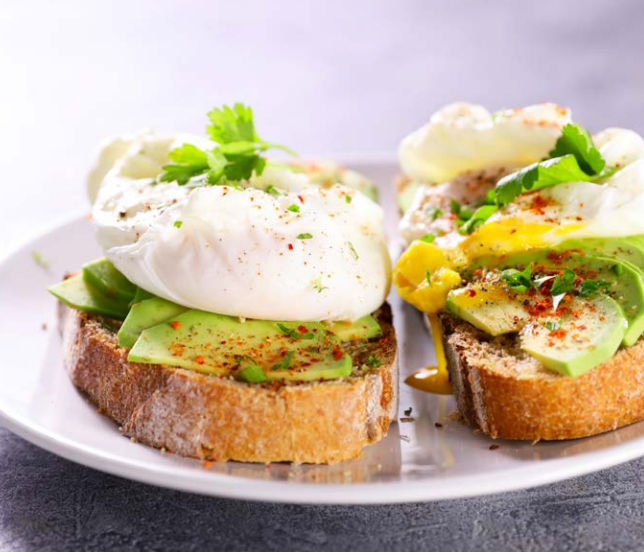 Sourdough toast topped with sliced avocado, poached eggs, fresh herbs, and a sprinkle of seasoning, served on a white plate.